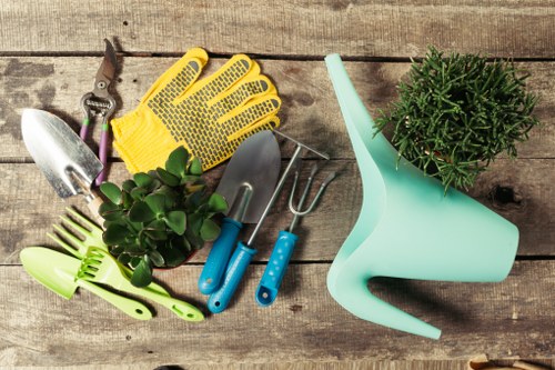 A collection of gardening tools and supplies arranged on a weathered wooden surface, including a pair of yellow rubber gloves, a small hand trowel and transplanter with metal blades and blue handles, a green plastic spray nozzle, a small potted shrub with green foliage, and a larger plastic watering can in light blue. The scene showcases typical outdoor gardening items used for planting, watering, and general garden maintenance, set in a natural outdoor environment with a rustic wooden backdrop. The arrangement suggests preparation for gardening tasks such as potting, soil tending, and watering, fitting the context of landscaping and garden care services offered by Landscapers Richmond in the Richmond area, with an emphasis on local gardening needs and outdoor maintenance in a professional setting.