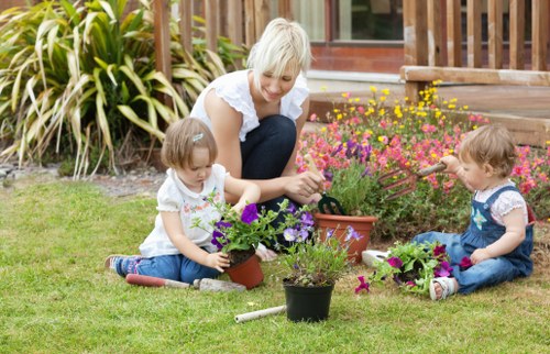 A woman with short blonde hair, dressed in a white top and dark jeans, is sitting on a well-maintained grassy lawn in a garden, accompanied by two young children. The children, a girl and a boy, are engaged in planting and tending potted plants, with the girl holding a purple-flowered plant and the boy reaching towards a pink-flowered plant with a gardening tool. Surrounding them are various flower pots filled with blooming flowers in shades of purple, pink, and yellow, which add vibrant colour to the scene. Behind them, there is a wooden deck with steps leading up to a house, and lush garden beds containing shrubs and ornamental plants, including tall grasses and flowering bushes. The garden appears to be well-kept, with neatly trimmed grass and an inviting outdoor environment suitable for family gardening activities, possibly part of a typical residential property in Richmond or nearby areas, reflecting local gardening interests and outdoor care practices facilitated by professionals like Landscapers Richmond. The scene takes place during daylight hours with natural lighting, creating a cheerful and productive outdoor setting.