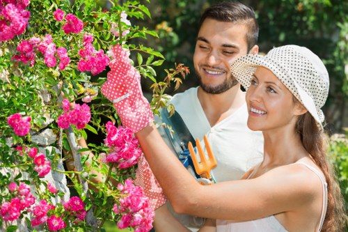 A couple working together in a lush garden area with vibrant pink flowering bushes and green foliage, positioned near a well-maintained lawn with dense grass and a defined flower bed border. The woman, wearing a white hat and gardening gloves, is holding pruning shears and appears to be pruning or tending to the flowering shrubs, while the man, dressed in casual clothing, observes and smiles. The background features trees and hedges, creating a natural outdoor setting suitable for gardening and landscaping services in Richmond. The weather is sunny with natural daylight illuminating the scene, emphasizing the lush greenery and bright blossoms, showcasing an organized garden space ideal for professional outdoor gardening work.