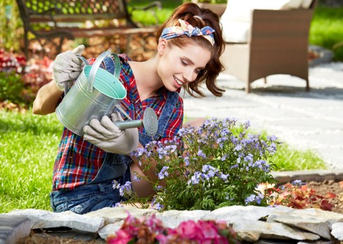 A young woman with light skin, wearing a plaid shirt, denim overalls, gardening gloves, and a colorful headband, kneels in a well-maintained garden in Richmond. She is tending to a small flowering bush with clusters of purple and light blue blooms, using a metal watering can to water the plants. The garden features a neatly edged flower bed with vibrant pink and purple flowers, bordered by natural stone edging. Behind her, there is a paved outdoor patio area with a white outdoor chair made of woven material, and a wooden garden fence surrounding the space. The scene is illuminated by natural daylight, suggesting a mild, sunny day, with lush green grass and some trees visible in the background. The overall environment appears tidy and cared for, reflecting the professional gardening services provided by Landscapers Richmond, enhancing outdoor spaces in the local area around Richmond.