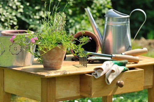 A wooden outdoor gardening table positioned in a lush, green garden setting, featuring various gardening tools and plant containers. On the table, there are several small terracotta pots with blooming pink flowers and green foliage, along with a larger, empty dark pot. A silver watering can with a long spout is placed towards the back of the table, adjacent to a pair of gardening shears, a garden fork, and a trowel with light-colored handles. A pair of gardening gloves is casually draped over the edge of the table, which appears to be used regularly for planting and tending to garden beds. The background displays a vibrant garden environment with dense greenery, including shrubs and trees, suggesting a well-maintained, cultivated outdoor space typical of residential gardens in Richmond or nearby areas. The scene is illuminated by natural daylight, with subtle shadows indicating a clear, mild weather day, supporting professional garden maintenance and landscaping services offered by Landscapers Richmond.