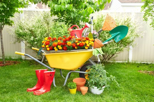 A vibrant backyard garden scene featuring a yellow wheelbarrow filled with colourful red and yellow flowers, positioned on a well-maintained green lawn. Surrounding the wheelbarrow are gardening tools such as a red watering can, a teal and orange small shovel, and a garden fork, indicating active maintenance. Nearby, there are small potted plants and empty terracotta plant pots placed on the lawn, with a white pot containing a lush green plant. In the background, a wooden fence provides privacy, with mature trees and bushes offering shade and natural foliage. The scene is illuminated by natural daylight, suggesting a mild weather day, typical of outdoor garden care in Richmond or nearby areas. This setting reflects standard gardening and landscaping activities carried out by local specialists like Landscapers Richmond, focusing on outdoor maintenance and garden enhancement.