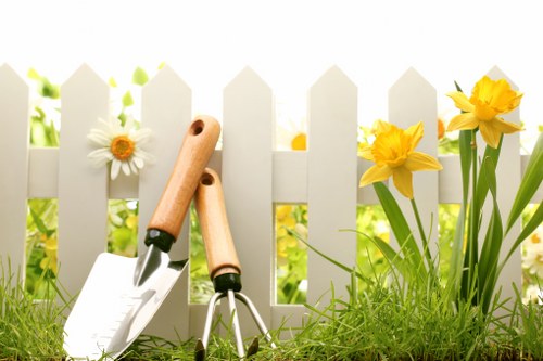 A close-up view of a small garden bed in a residential outdoor space in Richmond, featuring a white picket fence at the back. In the foreground, there is lush, green grass with a slightly uneven texture. To the right, bright yellow daffodils with elongated green leaves grow next to the fence, adding vibrant colour to the scene. Leaning against the fence are gardening tools, including a trowel with a wooden handle, a hand rake, and a pair of pruning shears, suggesting recent or ongoing gardening and lawn maintenance. The background has natural sunlight illuminating the scene, creating soft shadows and highlighting the fresh, healthy appearance of the plants. This image provides a clear depiction of a well-maintained garden area suitable for landscaping services focused on lawn care, flower bed upkeep, and outdoor space enhancement in Richmond.