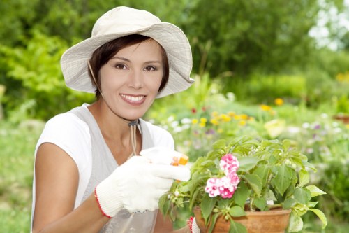 A woman wearing a wide-brimmed white sun hat and gardening gloves is tending to a potted flowering plant with pink and white blossoms in a lush outdoor garden. The garden features a well-maintained lawn with dense, green grass in the foreground, bordered by flower beds and shrubs, with a variety of plants and small trees visible in the background. The scene appears to be set during daytime with natural sunlight filtering through the foliage, creating a bright and inviting atmosphere. The woman is smiling and appears engaged in gardening tasks, contributing to the overall impression of outdoor maintenance and garden care that aligns with professional landscaping services provided by Landscapers Richmond. The environment reflects a healthy, vibrant garden space typical of residential or community gardens in the Richmond area, with attention to plant health and garden aesthetics.