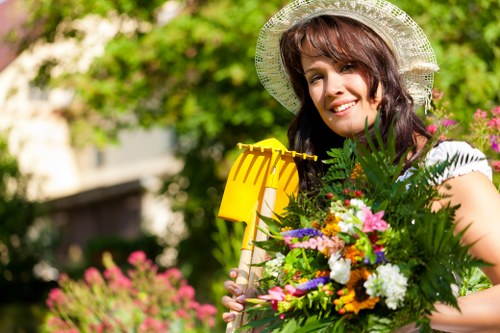 A woman with shoulder-length brown hair, wearing a white sunhat and a white top, is standing outdoors in a garden during daylight hours. She is smiling and holding a large colorful bouquet of various flowers and greenery in front of her. The garden background features lush green trees, some pink flowering plants, and a blurred garden hedge, suggesting a well-maintained outdoor space. The sunlight illuminates the scene softly, highlighting the vibrant colors of the flowers and the natural tones of the garden environment. This setting emphasizes aspects of outdoor gardening and landscaping, suitable for services such as garden design, planting, and landscape maintenance offered by landscapers in Richmond. The scene captures the essence of a vibrant, cultivated garden space, reflecting professional landscaping and gardening care in a local UK context.