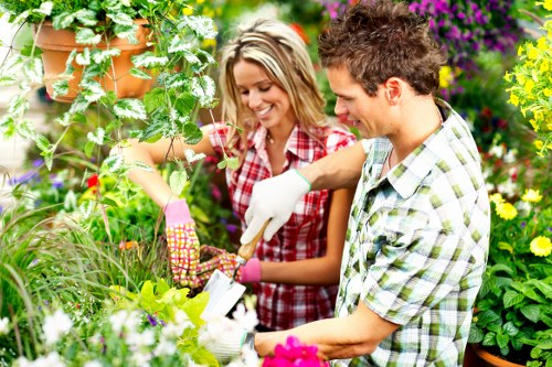 The image depicts two gardeners working together in a lush outdoor garden setting, with a variety of vibrant flowering plants and greenery. The foreground shows a well-maintained patch of grass and a flower bed with blooming flowers in shades of pink, yellow, and purple. Behind them, an array of shrubs and small trees with healthy green foliage create a natural backdrop, while hanging plant pots and climbing vines add layered texture to the scene. The garden appears to be part of a residential outdoor space, with a mix of soil and paved areas visible, and natural sunlight illuminating the scene, suggesting fair weather. The gardeners are smiling and engaged in planting or tending to the plants, highlighting elements of gardening and landscaping services, such as planting, soil management, and plant care, which could be offered by landscapers in Richmond or similar locations.