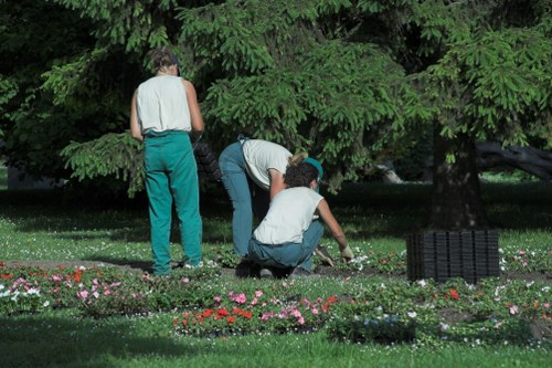 Three individuals are working together in a lush garden in Richmond, with dense, leafy green trees serving as a backdrop. The scene shows two women and one man engaging in garden maintenance near a flower bed containing small, colorful blooming plants, including pink, red, and white flowers, arranged along the edge of a well-kept lawn. The woman on the left is standing, wearing a sleeveless top and teal trousers, while the woman in the middle is bent over, tending to the plants, dressed in a light top and dark trousers, with a gardening glove on her right hand. The young man on the right is kneeling, actively planting or weeding, wearing a white shirt and dark trousers. The garden surface includes a combination of grass and soil patches, with a black garden debris basket visible on the right side. The overall scene reflects eco-friendly outdoor maintenance typical of high-quality local gardening services in the Richmond area, emphasizing natural textures and the diversity of yard plantings under natural daylight.