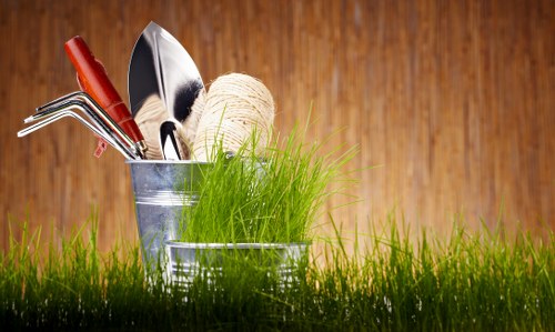 A close-up view of a small collection of gardening tools and supplies placed on a patch of vibrant green grass outdoors. The tools include a red-handled garden fork and a trowel with a stainless steel blade, alongside a spool of natural twine and a small pot of bright green grass or young plants. The background features a wooden surface, suggesting a garden shed or fence, with warm natural lighting indicating daylight conditions. The arrangement highlights typical gardening equipment used for planting, maintenance, and outdoor landscaping, aligning with professional gardening and landscaping services offered by companies like landscapersrichmond.co.uk in Richmond or nearby areas, and emphasizing the importance of proper outdoor care and safety.