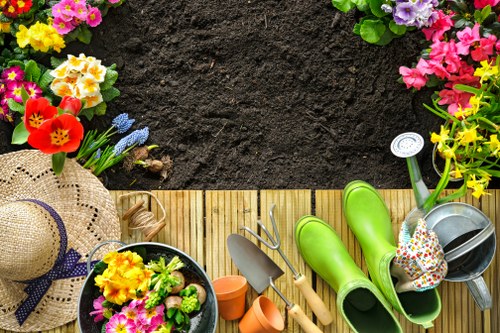 The image shows a section of a garden with a wooden decking area bordered by colorful flowering plants including pink, yellow, and purple blossoms, alongside green foliage. In the foreground, gardening tools such as a small trowel, fork, and watering can are neatly arranged on the decking, indicating recent or upcoming outdoor maintenance activities. A straw sunhat with a purple ribbon rests on the decking, suggesting a gardener preparing for work. Adjacent to the deck is a patch of dark, well-turned soil, possibly prepared for planting or awaiting further landscaping. The environment appears to be a well-maintained back garden, with natural lighting suggesting a bright, partly cloudy day, typical of outdoor spaces in Richmond. The combination of vibrant flowers, garden tools, and clean paving highlights the ongoing gardening and landscaping efforts undertaken by professional landscapers like Landscapers Richmond, focused on enhancing outdoor garden spaces in the local area, including the postcode RH.