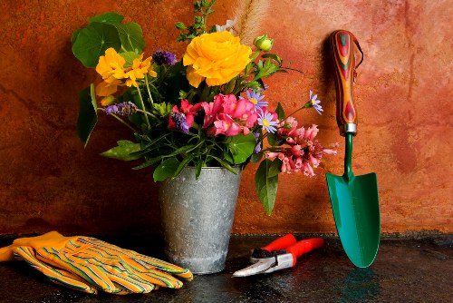 A still life image featuring a large, grey, cylindrical flower pot placed on a dark work surface, containing a vibrant arrangement of fresh flowers including yellow, pink, purple, and blue blooms with green foliage. To the right of the pot, gardening tools are arranged: a small green-handled trowel with a reddish-brown handle, a pair of red pruning shears, and a pair of gardening gloves with yellow and black stripes. The background is a textured, warm-toned orange wall that complements the natural colours of the flowers and tools, suggesting an outdoor or garden workshop environment typically associated with residential gardening and landscaping services in Richmond, postcodes within southwest London. The overall scene conveys tools and elements useful for garden maintenance, planting, and flower arrangement, aligning with professional gardening and landscaping practices in the local area.