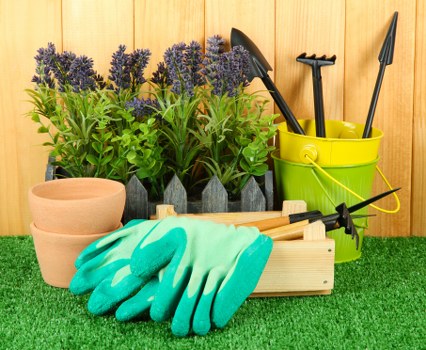 A collection of gardening tools and supplies arranged on a patch of artificial green grass in an outdoor setting. In the foreground, there are a pair of light blue rubber gloves placed on a wooden toolbox, which also contains small gardening hand tools. Behind the gloves, three terracotta plant pots are stacked together, with the top pot slightly tilted. To the left of the pots, several potted lavender plants with purple flower spikes and green foliage are visible, positioned in black containers. To the right, a bright yellow plastic bucket holds three gardening tools — a trowel, a transplanter, and a cultivator — all with black handles. The background features a wooden fence or paneling, indicating a garden or backyard environment, with natural lighting suggesting a clear day. The arrangement highlights essential garden maintenance tools and plant care supplies typically used for landscaping or gardening services in Richmond or similar UK locations, supporting professional outdoor garden upkeep activities.